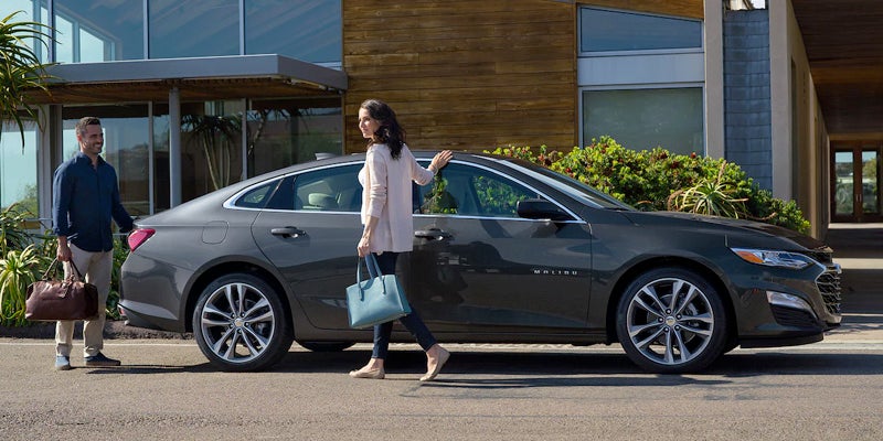 A man and woman with bags standing outside a dark gray sedan, parked in front of a modern house.