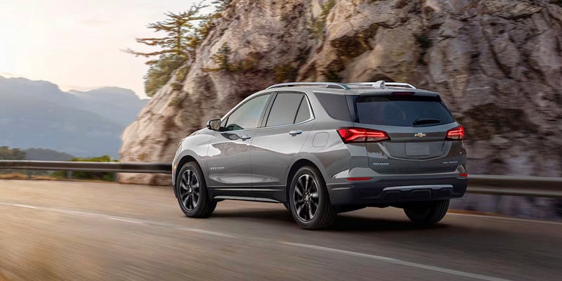 Rear three-quarter view of a gray Chevrolet Equinox SUV driving on a mountain road with sun-drenched hills.