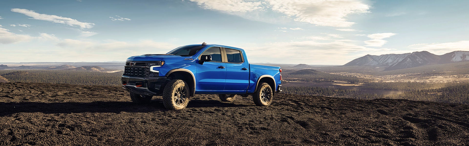 A blue Chevy Silverado AT4X truck parked on a dark, rocky hill with a forest and mountains in the background.