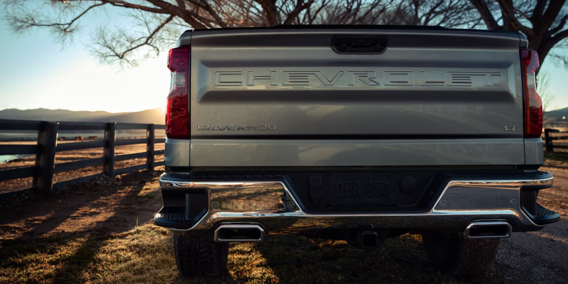 Rear view of a light gray Chevrolet Silverado truck with Chrome bumper, parked facing a sunset over mountains.