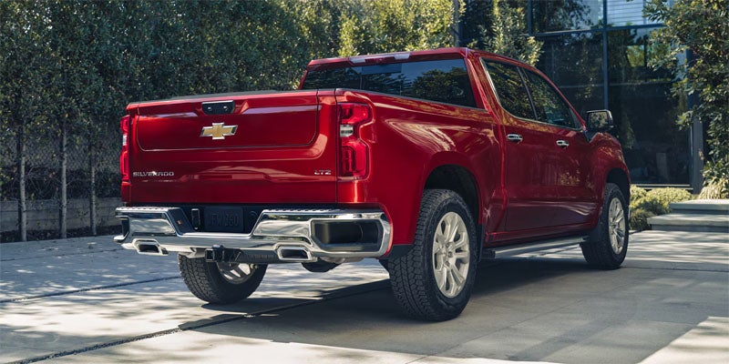 Rear view of a red Chevy Silverado pickup truck parked on a concrete driveway with green bushes and house in background.