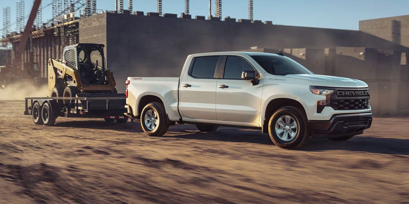 A white Chevrolet Silverado pickup truck towing a yellow skid-steer loader on a trailer at a dusty construction site.