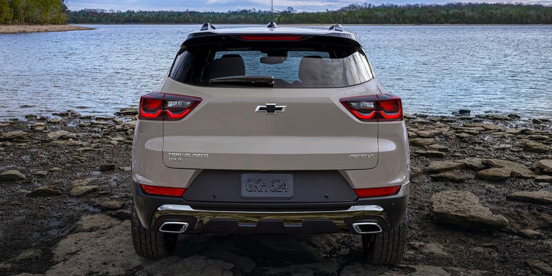 Front view of a light brown Chevrolet Trailblazer parked on a rocky shoreline next to a lake and trees.