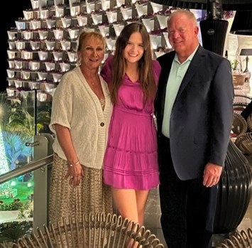 Young woman in pink dress stands between smiling older man in suit and older woman in cardigan against modern decor.