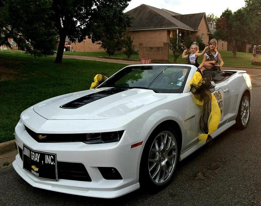 Two young girls with tiaras sit in the back of a white Chevy Camaro convertible decorated with yellow and black ribbons.