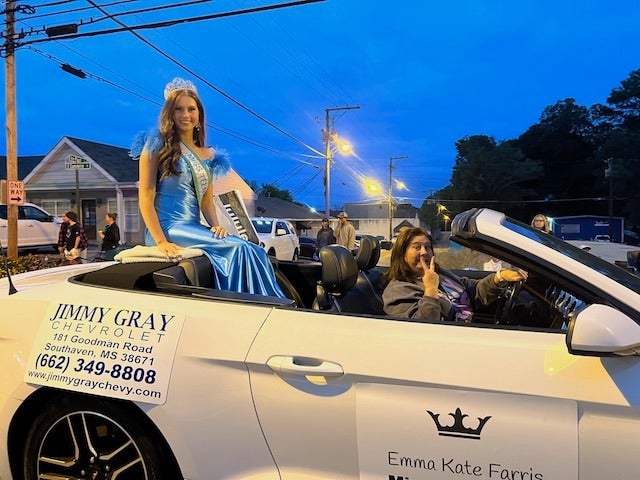 A pageant queen in a blue dress and crown rides in the back of a convertible with a driver, at dusk.