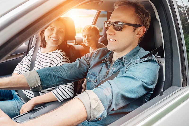 A family of three smiles while sitting inside a car during sunset